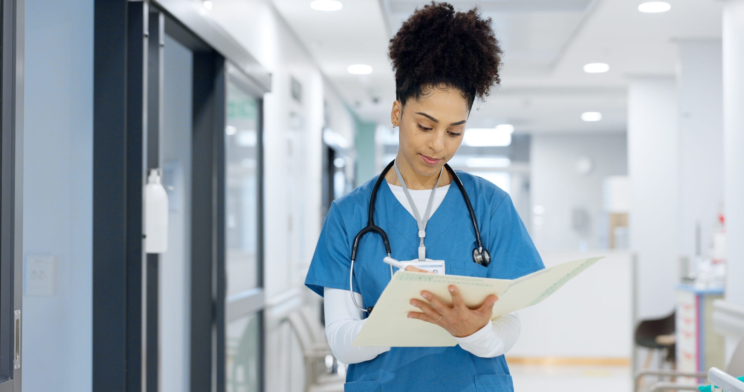 A nurse in blue scrubs, wearing a stethoscope around her neck, stands in a hospital hallway reviewing a patient's file.