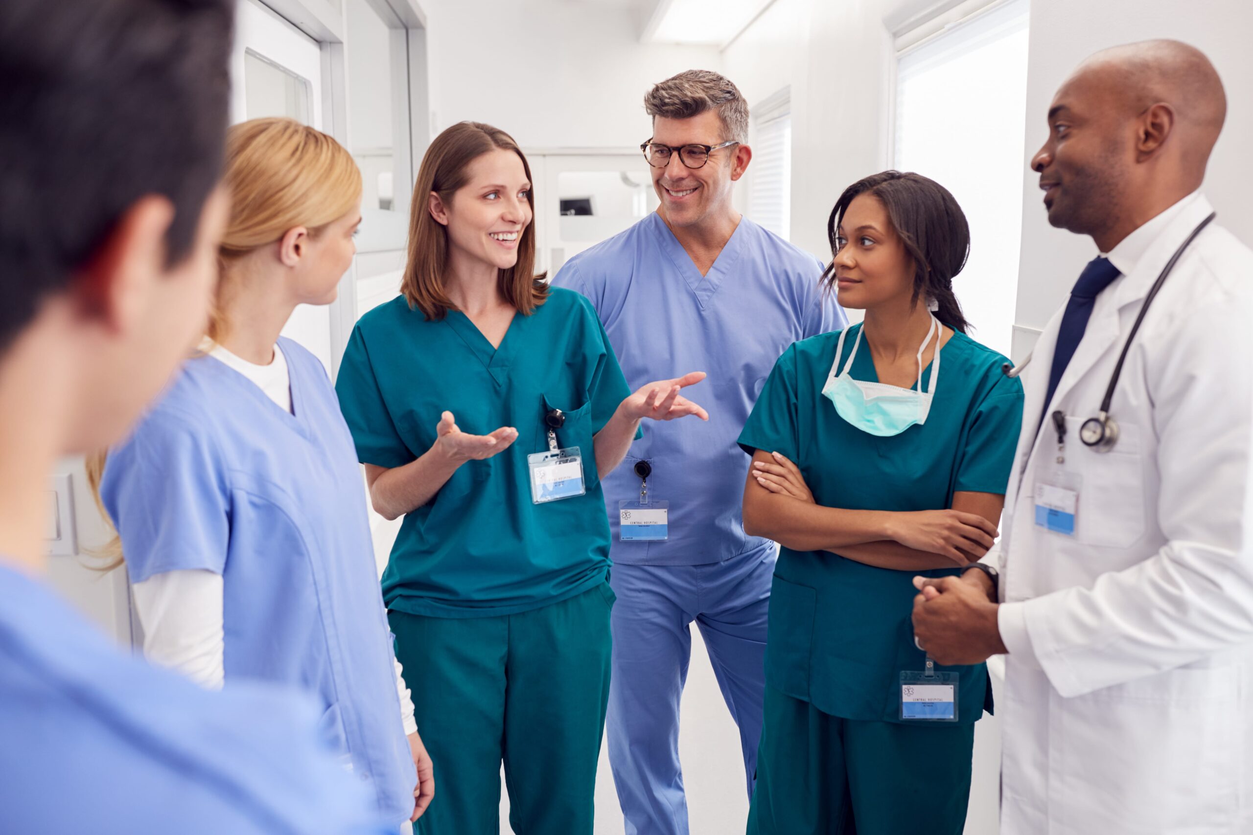 Healthcare team in scrubs and lab coats talking and smiling in a hospital hallway.