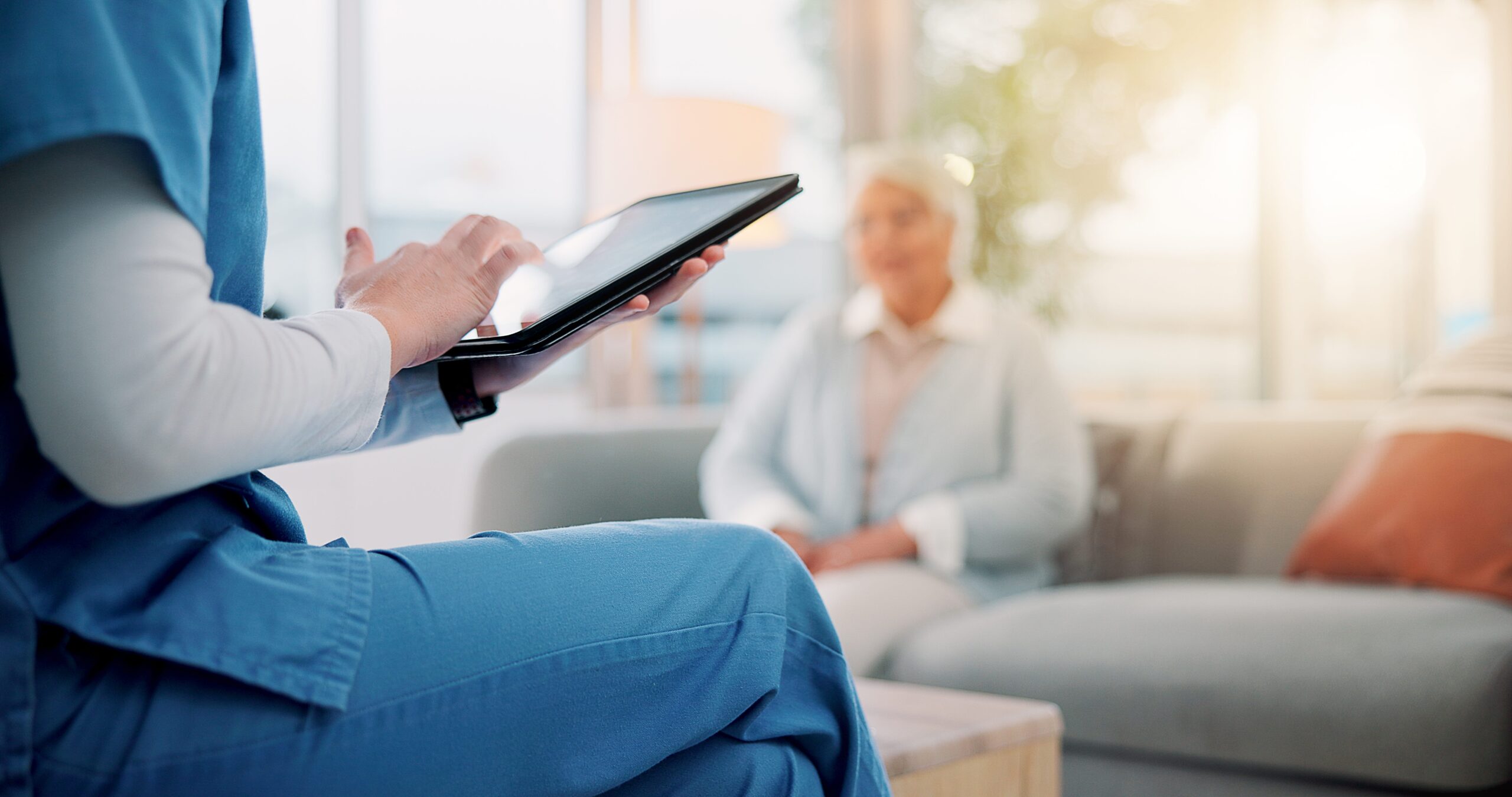 Nurse in scrubs using a tablet with a senior patient sitting on a couch in the background.