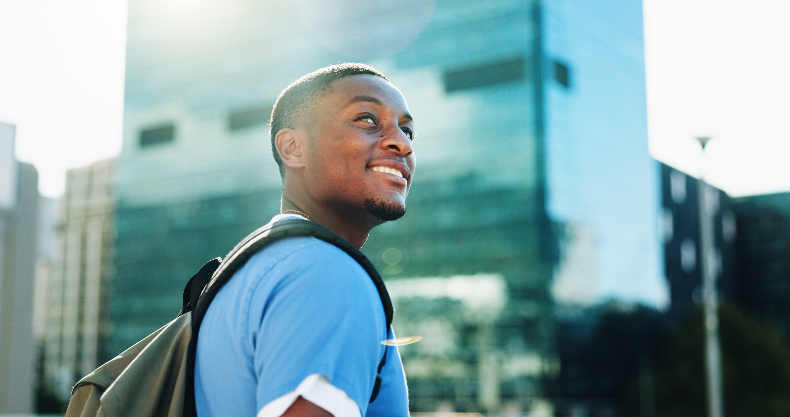Nursing student in blue scrubs with a backpack smiling outside a glass medical building.