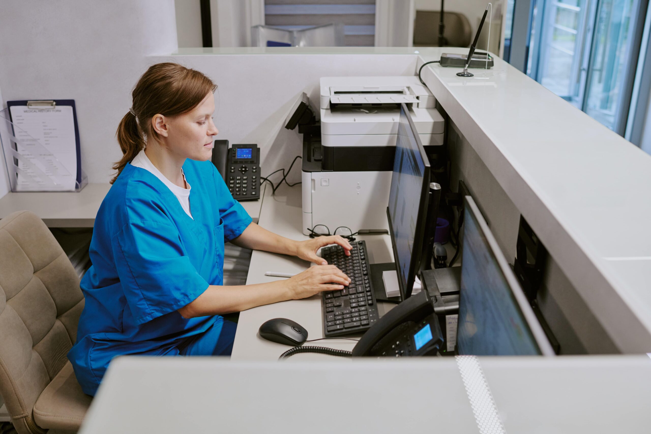 Nurse in blue scrubs working at a medical front desk, using AI to streamline patient documentation and digital records.