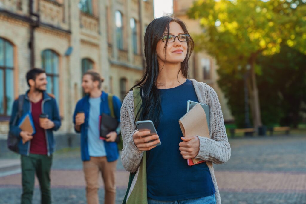 Confident female college student holding notebooks and a smartphone on campus, preparing to apply for an RN to BSN program.