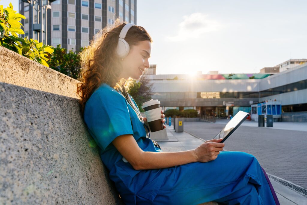 Nursing student in scrubs studying on a tablet outdoors while wearing headphones and holding a coffee cup.