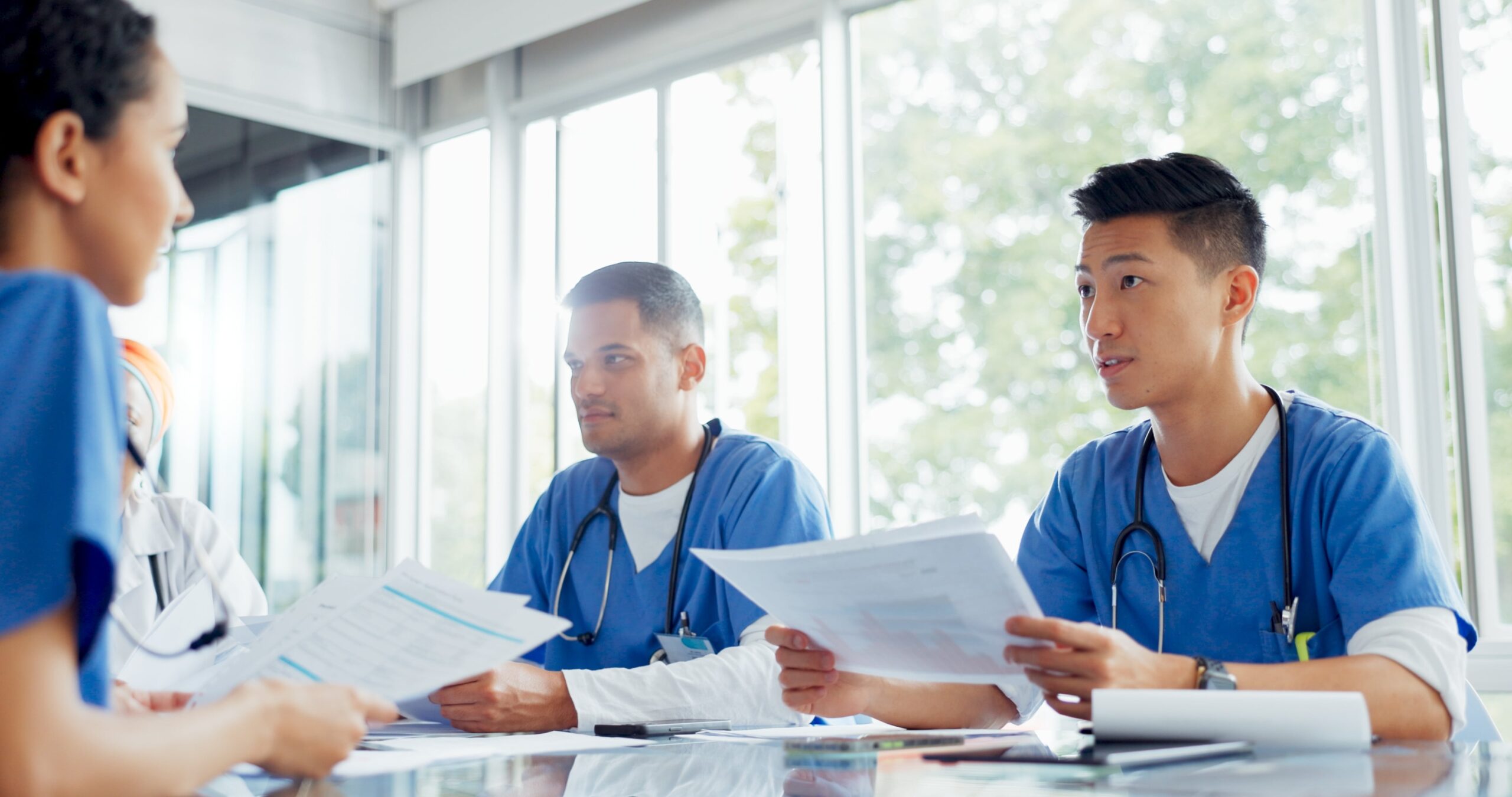 Group of nursing students in blue scrubs reviewing papers together in a bright classroom setting.