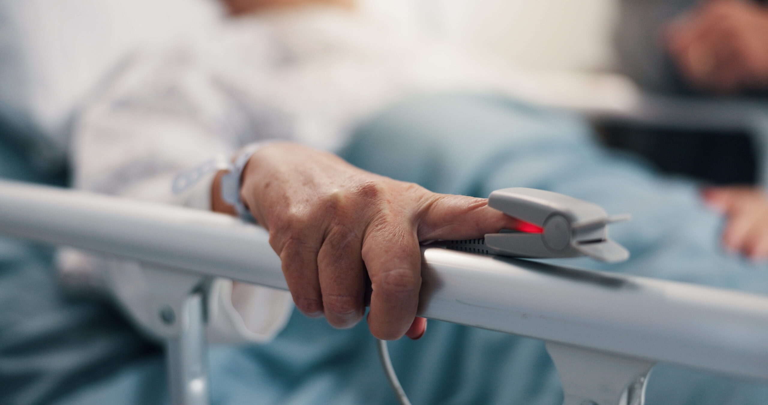 Patient’s hand resting on a hospital bed rail with a pulse oximeter attached.