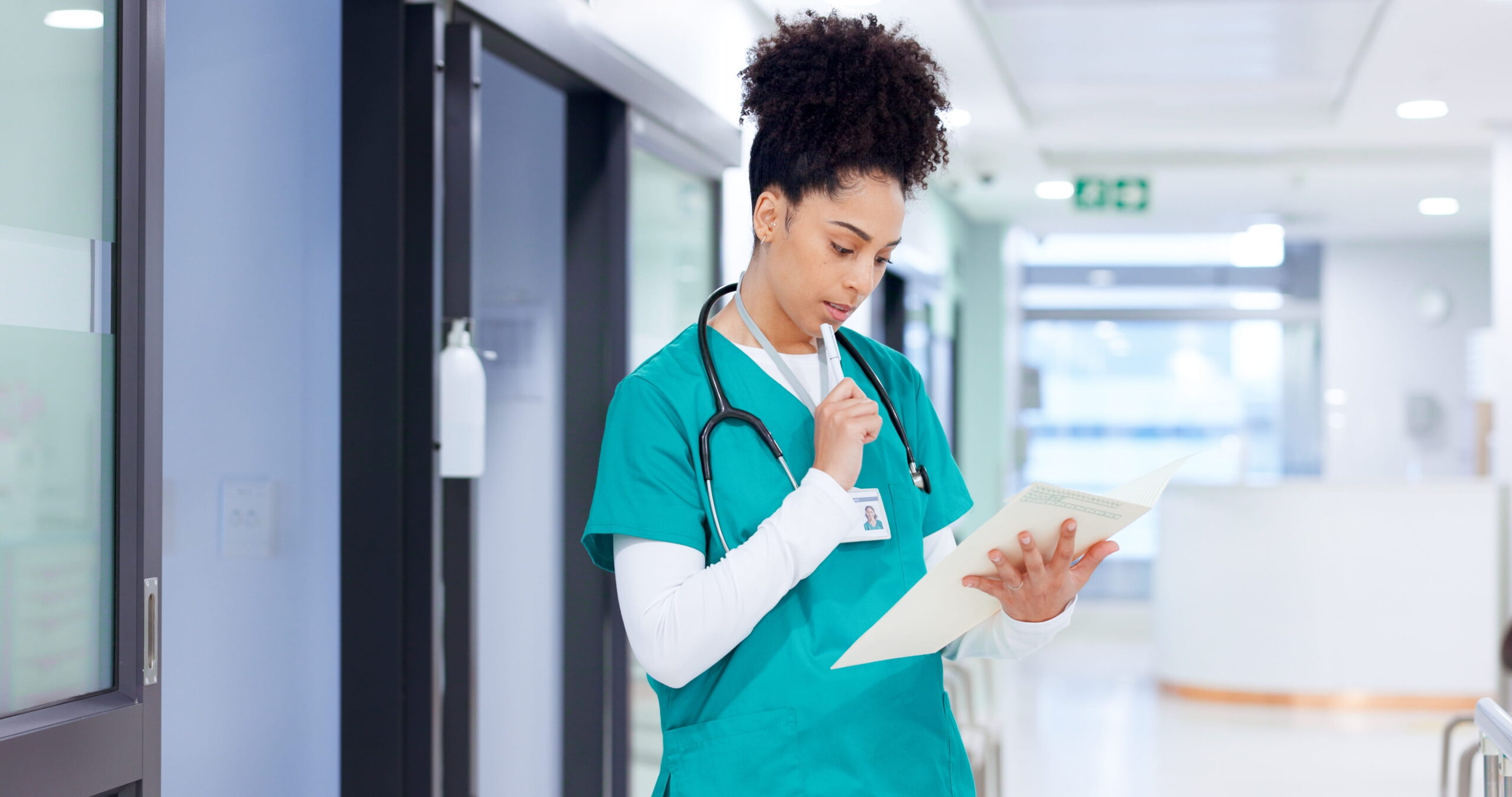Nurse in green scrubs reviewing a document and taking notes in a hospital hallway.