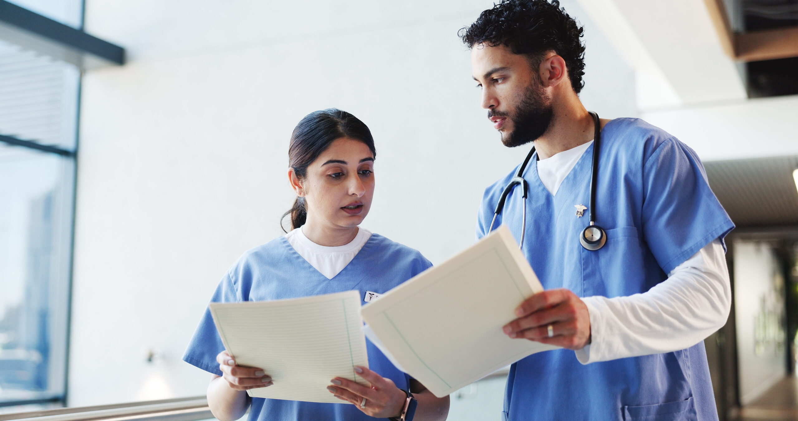 Two nurses in scrubs reviewing paperwork together in a hospital hallway.