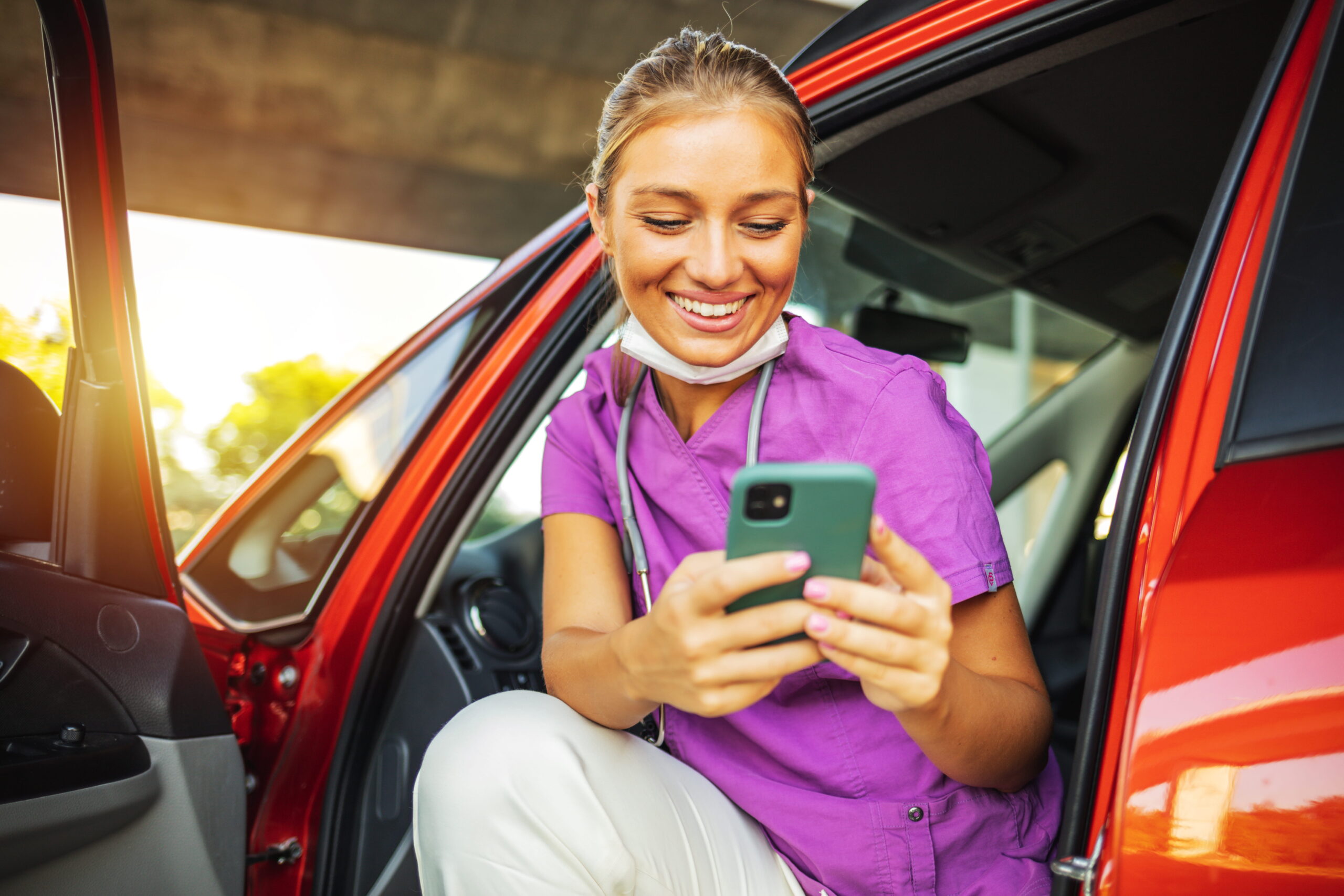 Travel nurse in scrubs sitting in a car doorway and looking at her phone.
