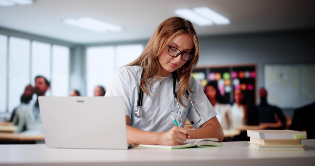 Nursing student studying course materials and taking notes beside a laptop in a classroom setting.