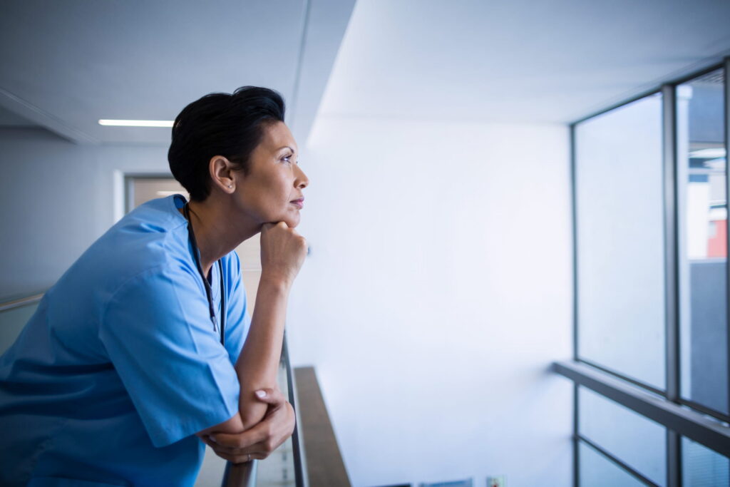 Nurse standing by a hospital window, thoughtfully looking out into the distance.