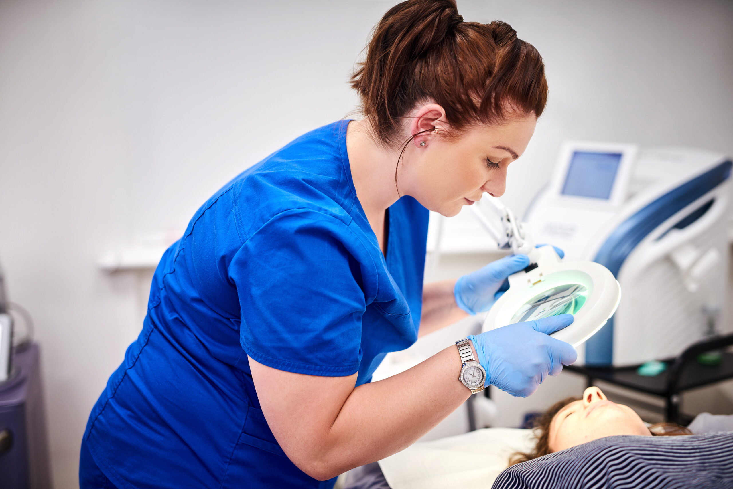 Aesthetic nurse examines a patient using a magnifying lamp during a cosmetic treatment.