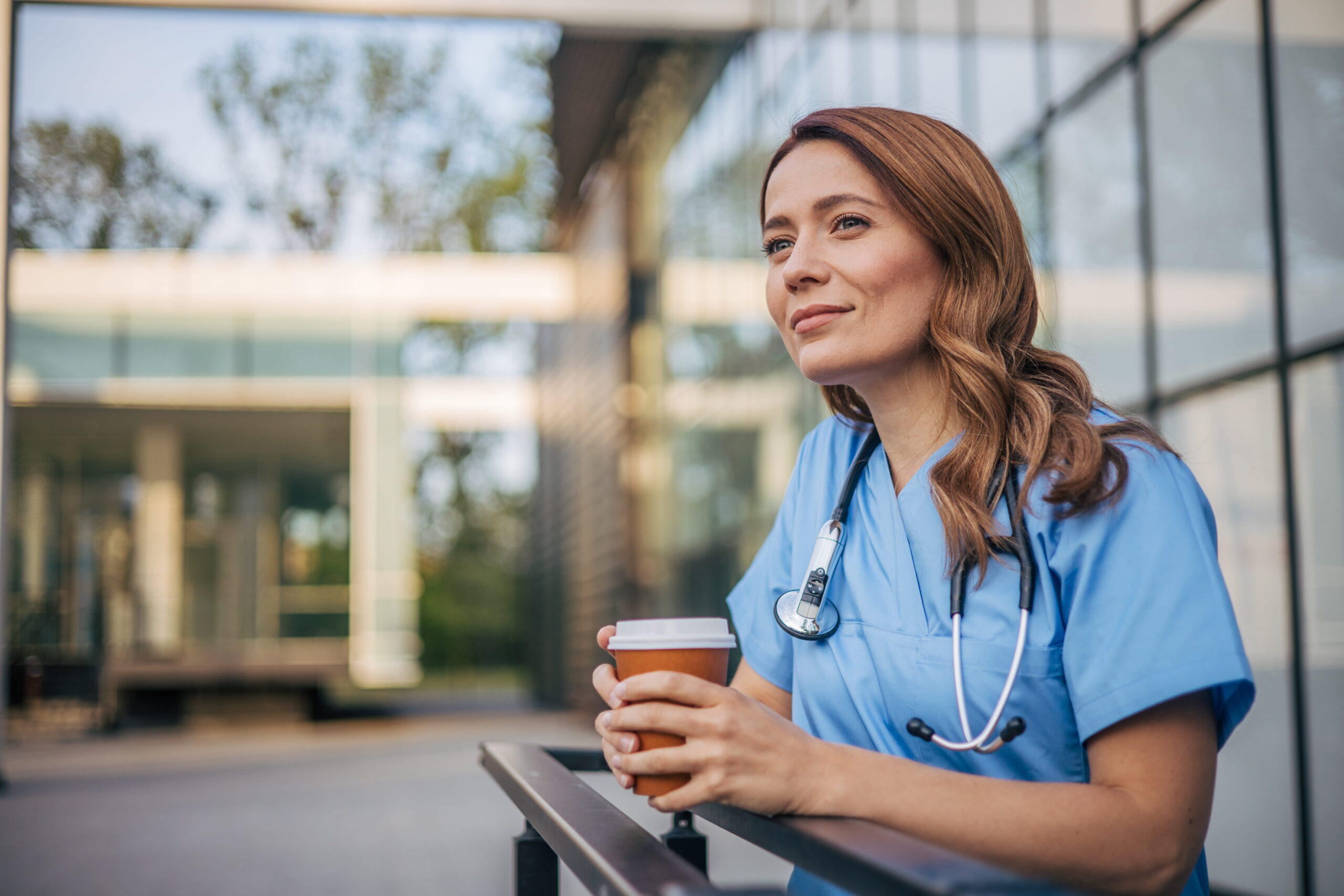 Nurse with a stethoscope holds a drink while taking a break outside a healthcare facility.